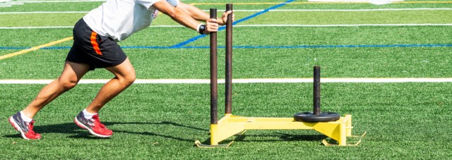 A high school teenage boy is pushing a yellow sled that has a 25 pound wieght on it during strength and conditioning practice.
