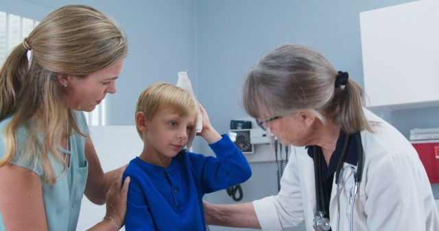 Child at pediatricians office holding ice pack on his head with mother and doctor in room. Little boy using cold compress in medical exam room receiving first aid