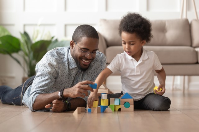 Caring young single black father help cute kid son play on warm floor together